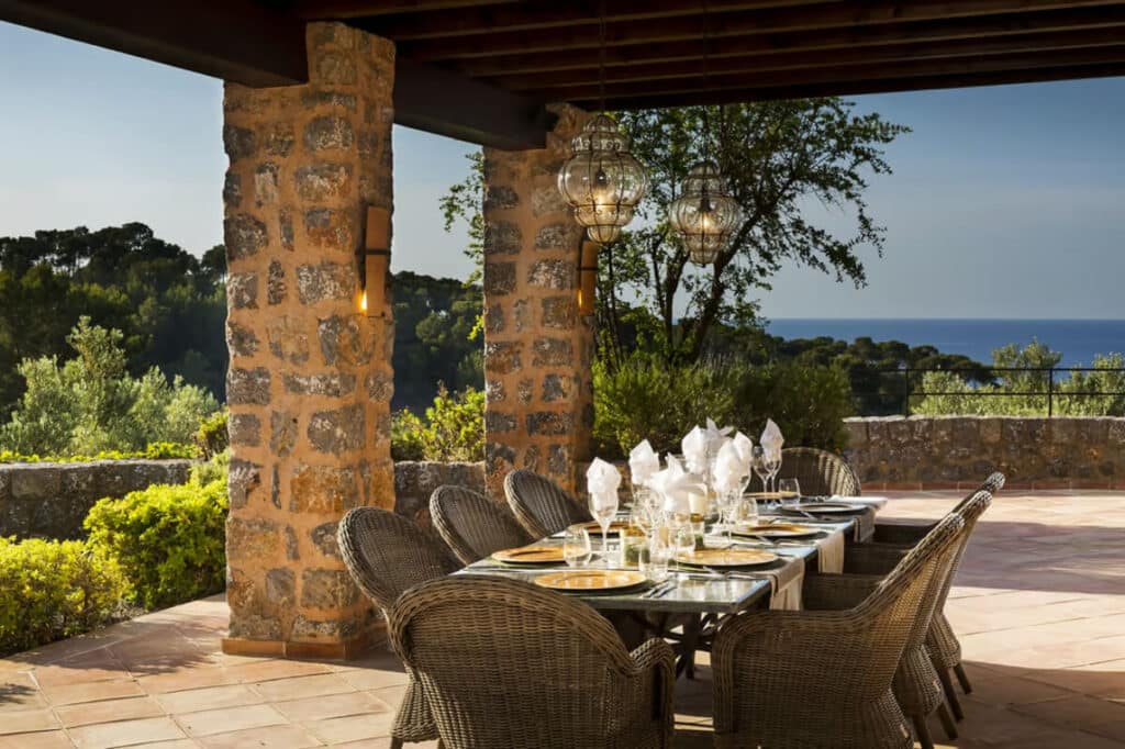 Elegant outdoor dining area under a rustic pergola with wicker chairs, hanging lanterns, and scenic views of greenery and the sea