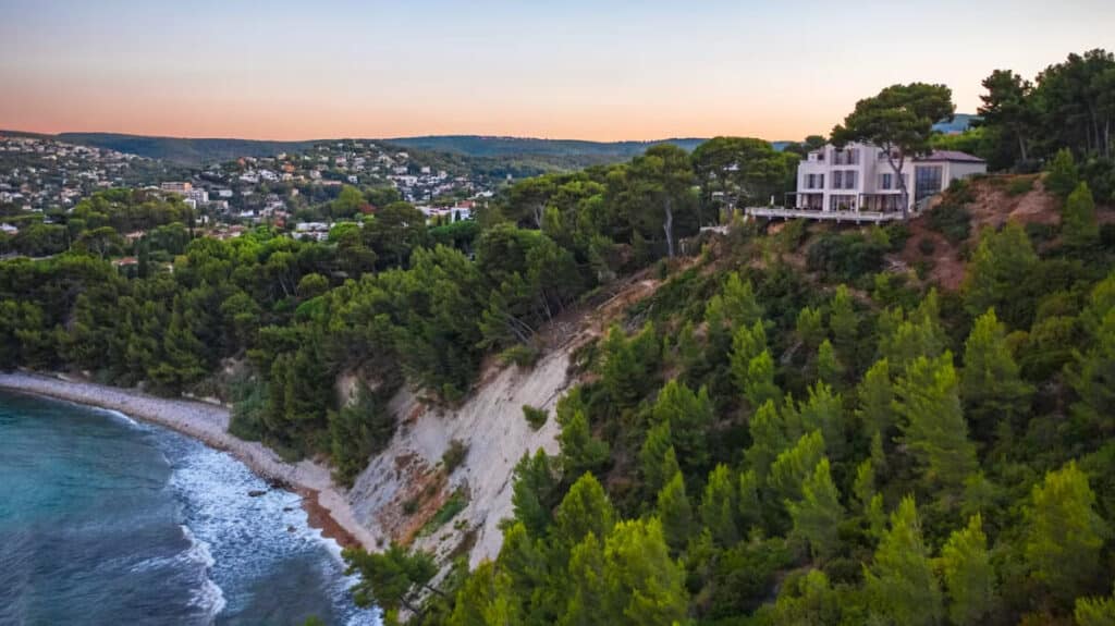 Modern villa atop a forested cliff overlooking a coastline at sunset