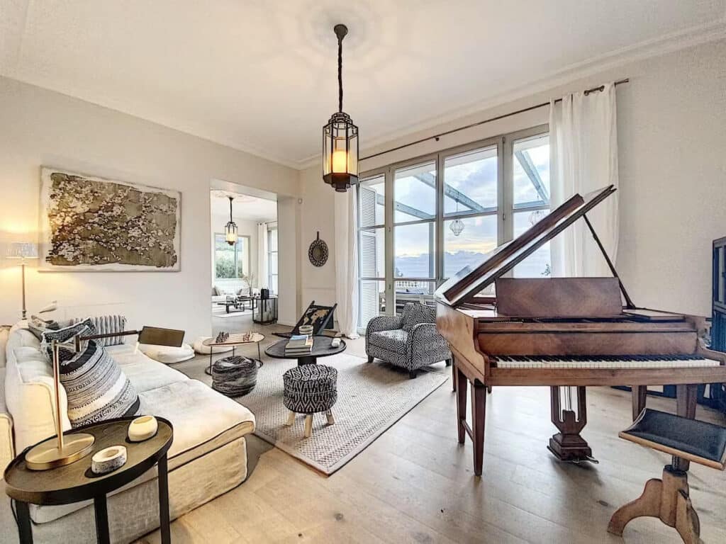 Elegant living room with a grand piano, large windows showcasing mountain views, cozy seating, and modern neutral-toned décor