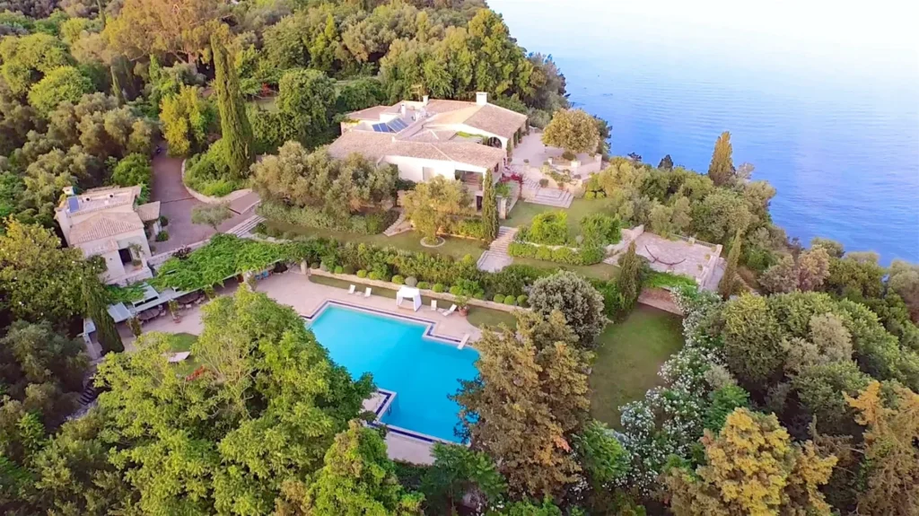 Aerial view of a hillside estate with large pool, terraced gardens and dense trees beside the sea.