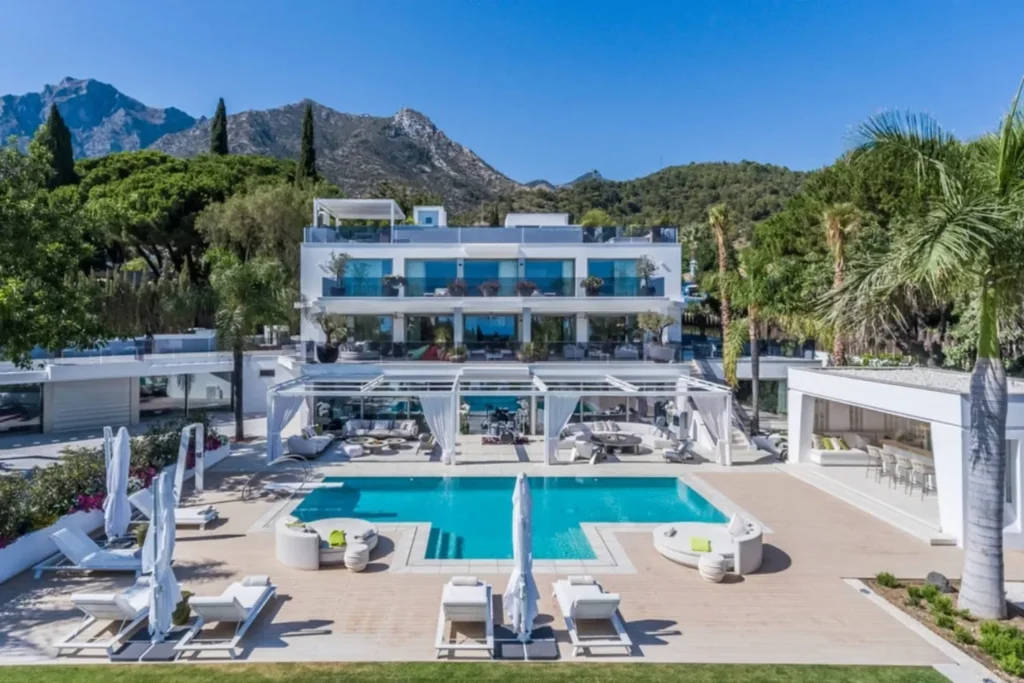 Contemporary white villa with tiered terraces, central pool, sun loungers and mountain backdrop.