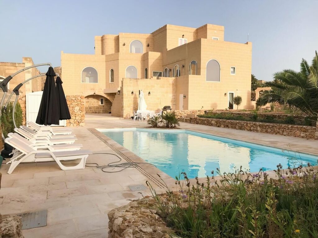 Poolside view of All That Glitters Is Gozo, a honey-stone farmhouse villa with sun loungers and palm trees.