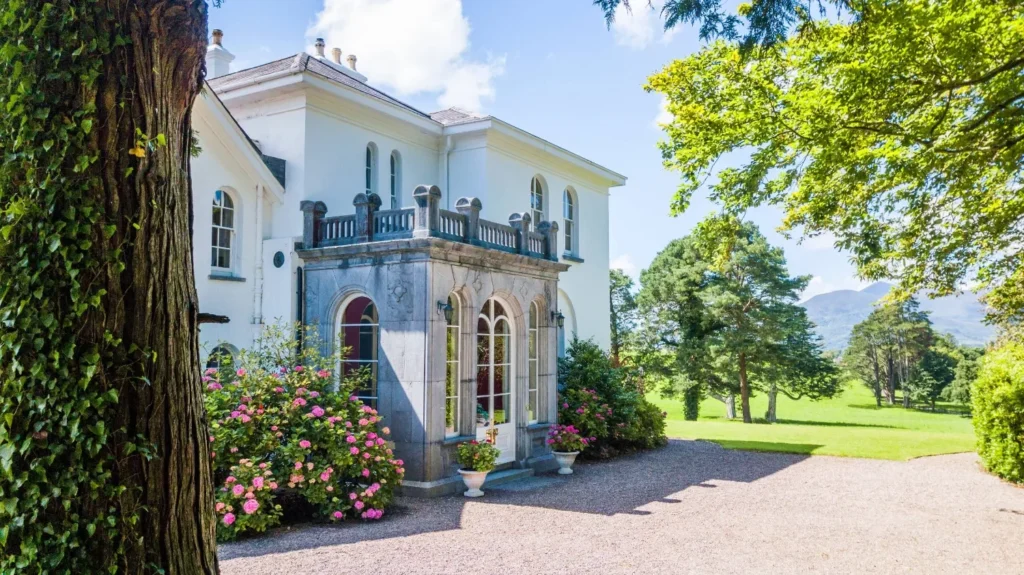 Elegant white manor house with a stone entrance and arched windows, framed by flowering shrubs, a gravel drive, and green lawns.