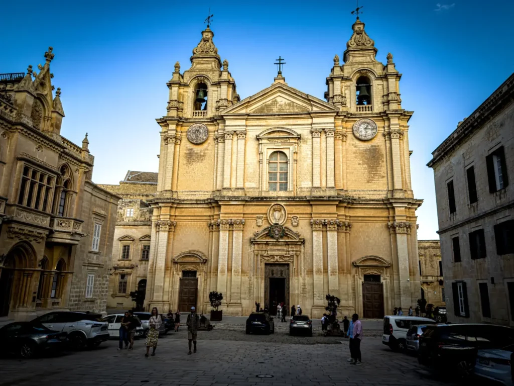 A grand sandstone church in Malta with twin bell towers and people gathered in the square out front.