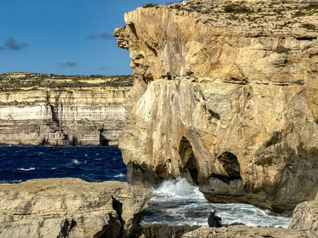 Person sitting on rocks beneath towering cliffs in Gozo’s Dwejra Bay as waves crash below.