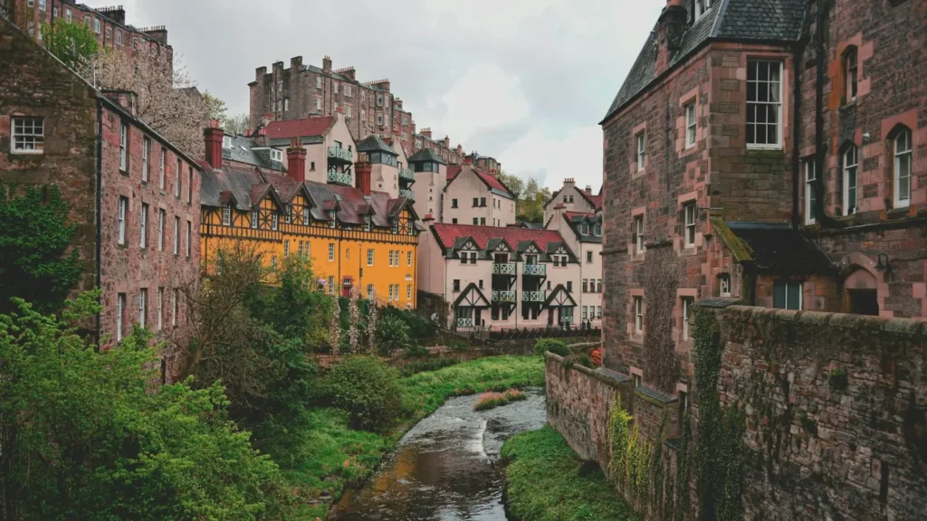 Colorful historic buildings in Edinburgh overlooking a narrow stream, with stone walls and greenery under a cloudy sky.