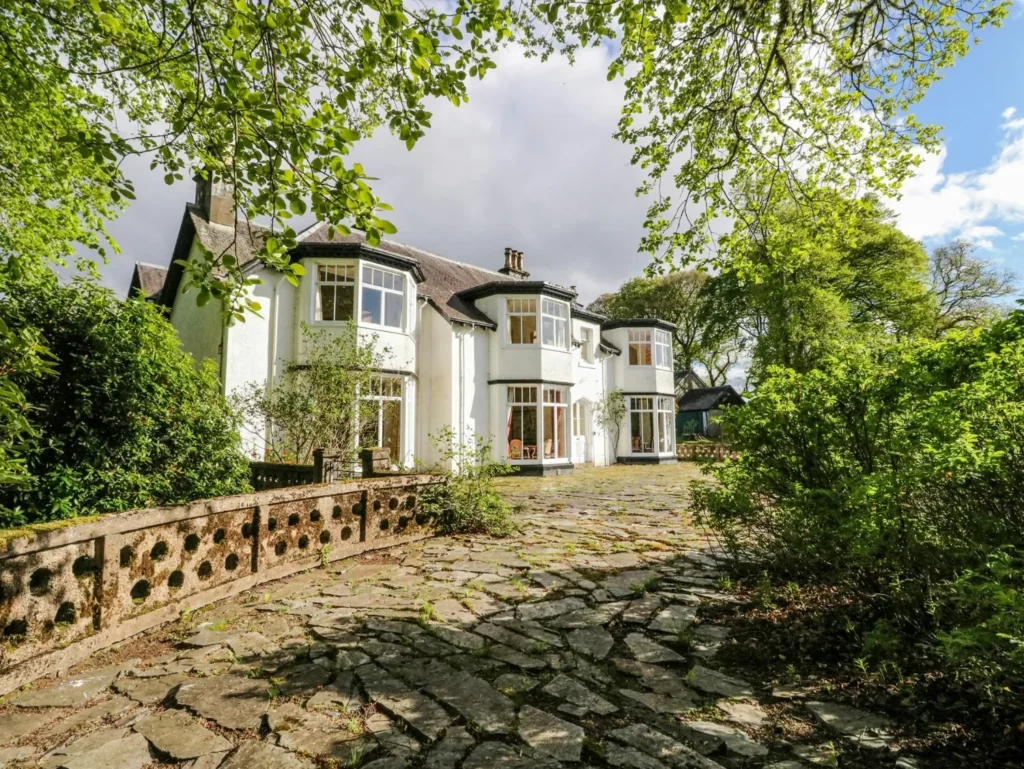 Large white country house with bay windows, set among lush trees and shrubs, with a wide stone-paved courtyard.