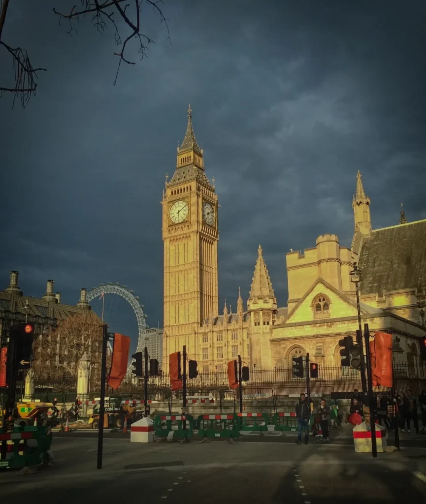Big Ben and the Houses of Parliament lit by low sun under dark clouds, with the London Eye in the background.