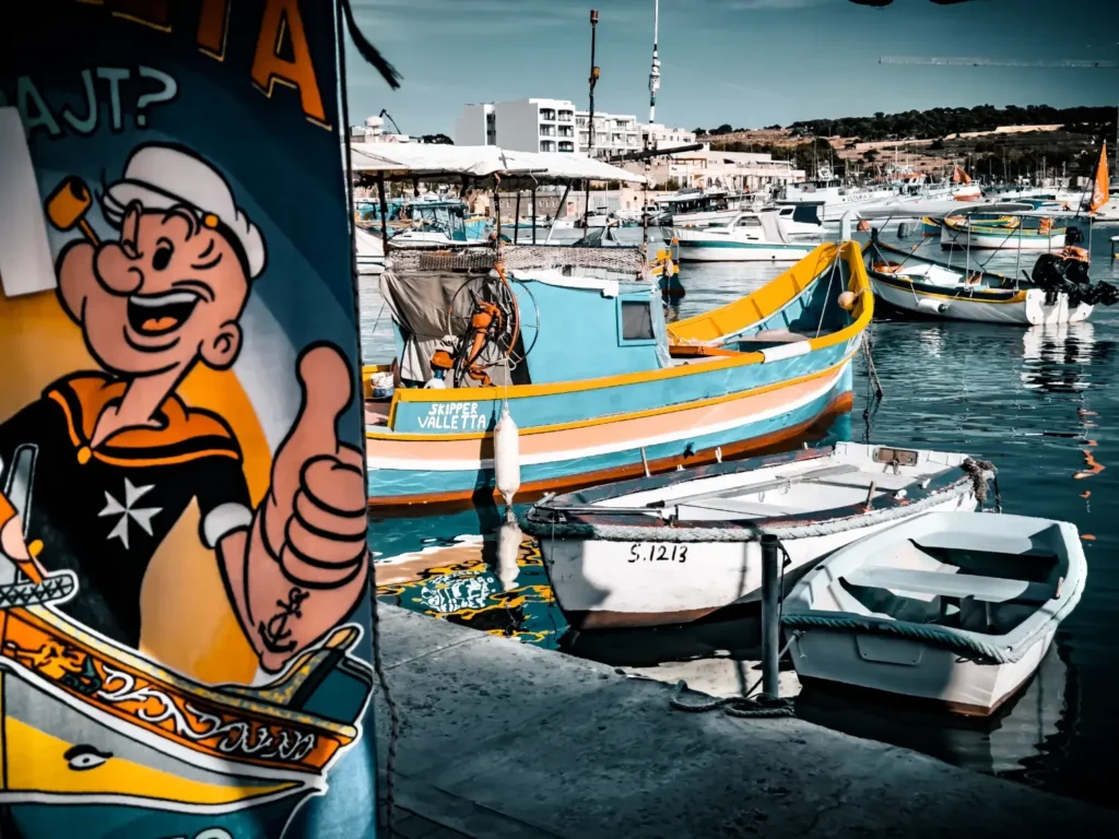 Colorful fishing boats in Malta’s Marsaxlokk harbor with a Popeye mural in the foreground.