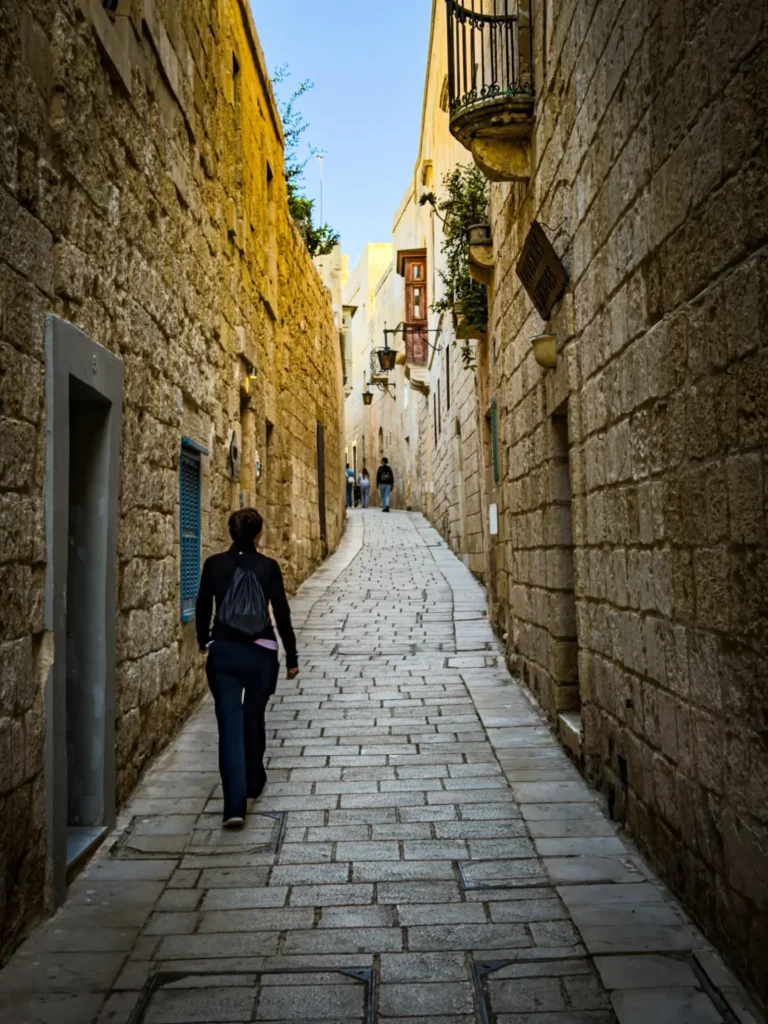 A tourist walking up a narrow stone alleyway between high honey-colored walls in Mdina, Malta.