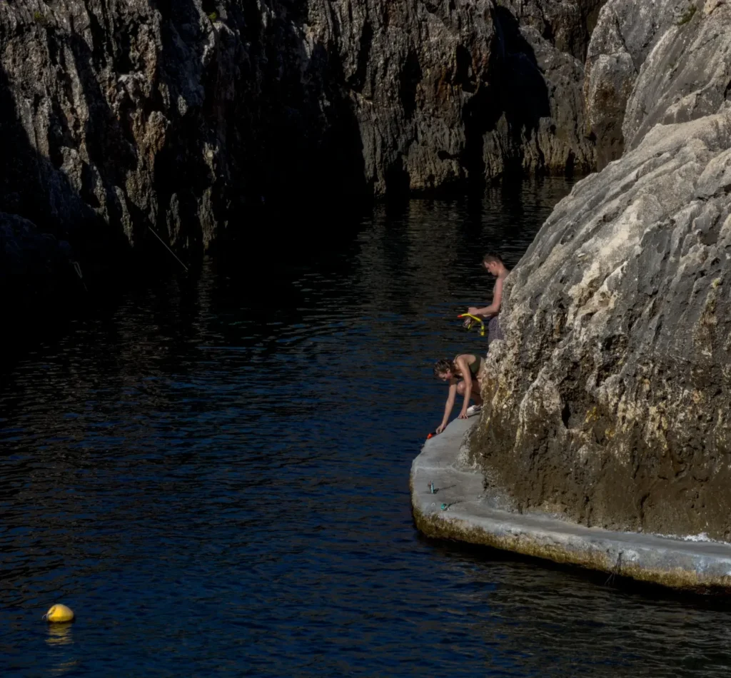 Two swimmers on a rocky ledge, preparing to snorkel in deep blue water near Malta’s Blue Grotto.