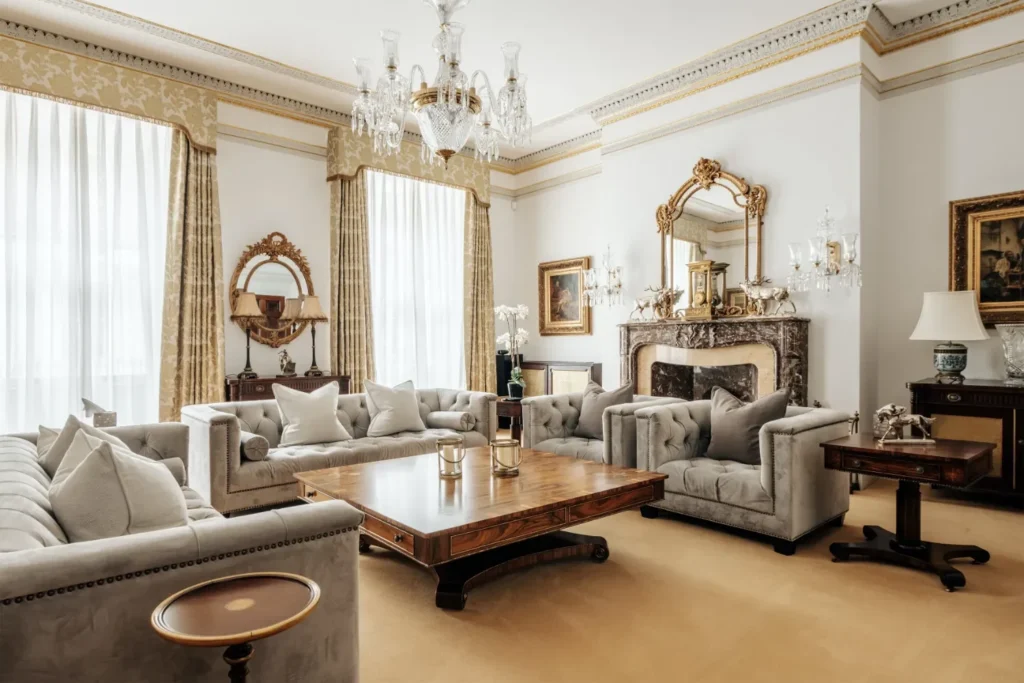 Elegant living room with grey tufted sofas, marble fireplace, gold mirror, chandeliers, and tall windows.