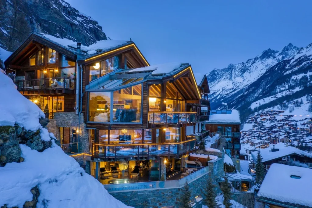Warm-lit glass-fronted chalet on a snowy mountainside overlooking a winter village and snow-capped peaks.