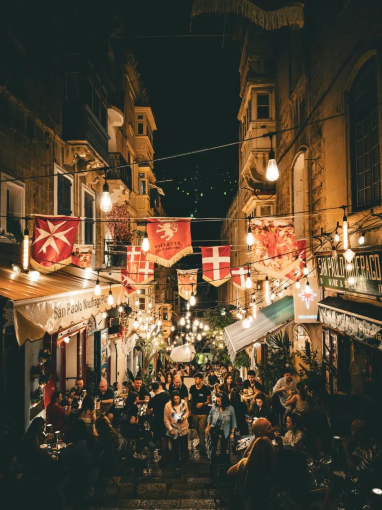In Malta, a crowded Valletta Street at night with lights, flags and people dining and celebrating outdoors.