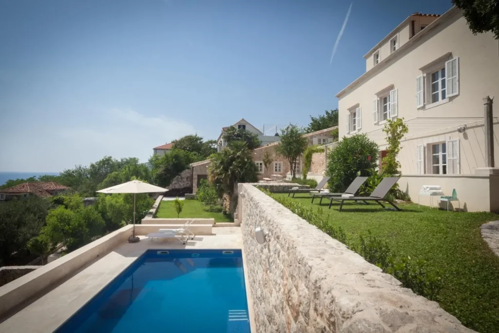 Cream hillside villa with private pool beside a stone wall, with sun loungers on a lawn.