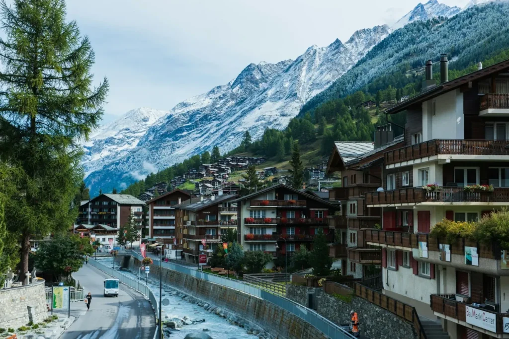 Chalet-lined street in Zermatt village with a shuttle bus and snowy mountains in the background.