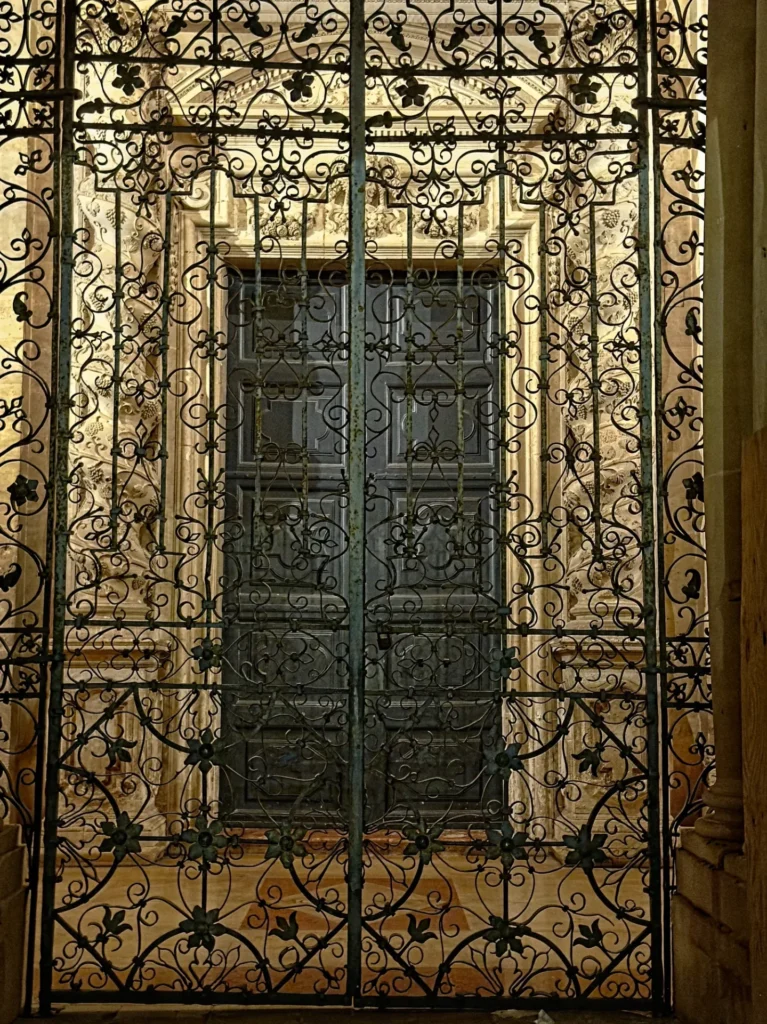 Original photo by paul partridge of an ornate wrought-iron gate framing a carved stone doorway of the cathedral of syracuse in ortigia, sicily.