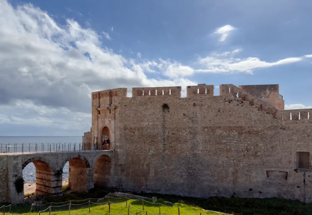 Original photo by paul partridge of the stone walls and arched bridge of castello maniace on ortigia’s seafront, silhouetted against a bright sky.