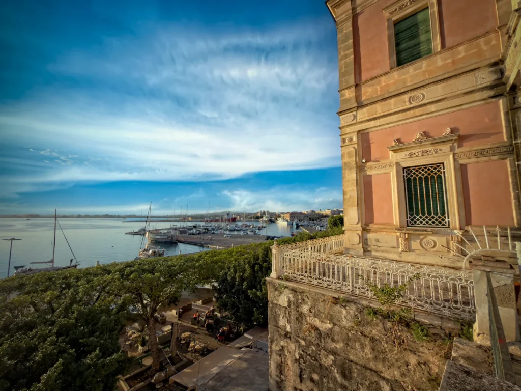 Original photo by paul partridge of ortigia’s harbor and the public gardens from the foro vittorio emanuele ii, with sailboats in the marina and a historic building façade on the right.
