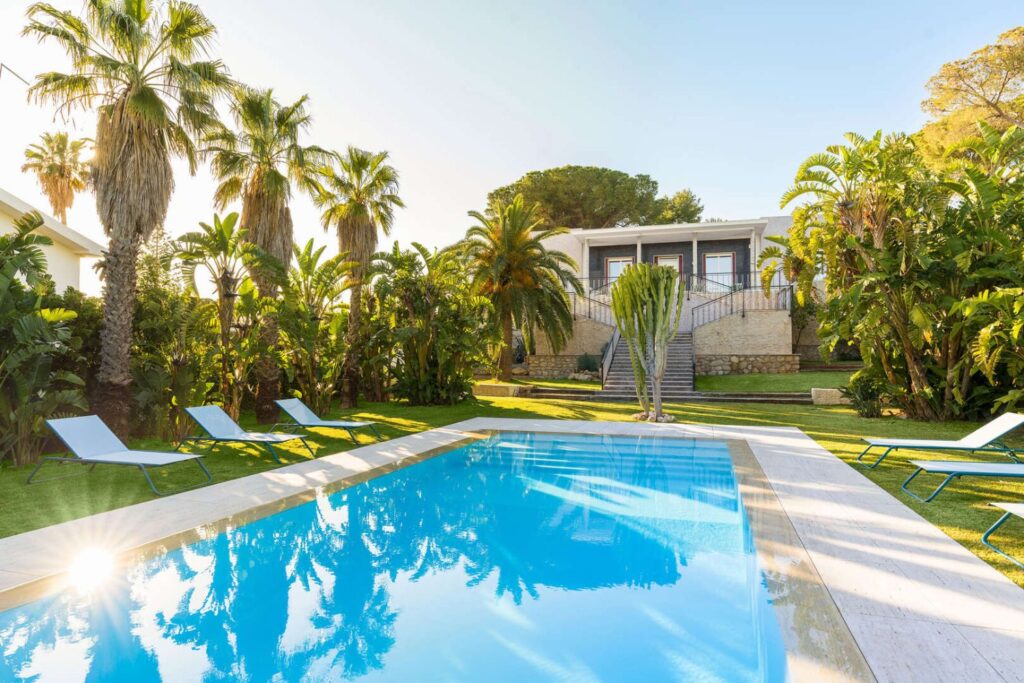 Swimming pool at la belle in fontane bianche, syracuse, surrounded by palm-filled gardens and sun loungers, with the villa set back on a raised terrace.