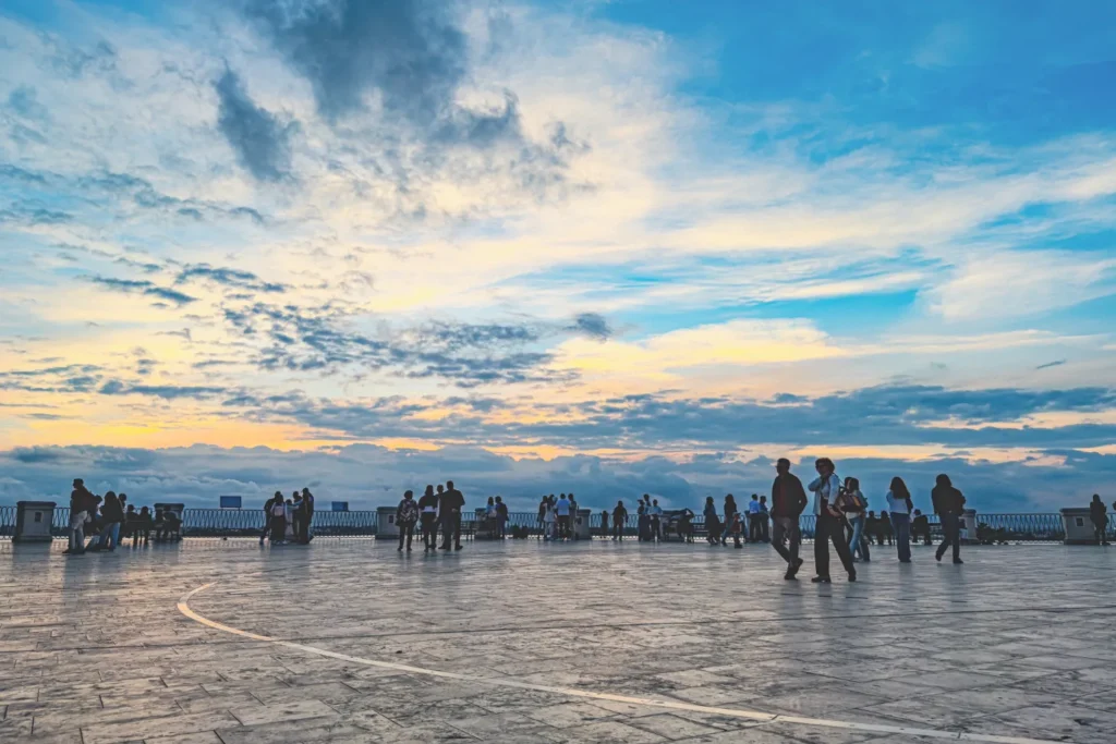 Original photo by paul partridge of people strolling along ortigia’s waterfront promenade at sunset near aretusa garden, silhouetted against a pastel sky.