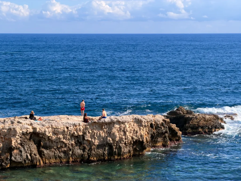 Original photo by paul partridge of sunbathers relaxing on rocky seaside cliffs in ortigia, with the deep blue ionian sea stretching to the horizon.