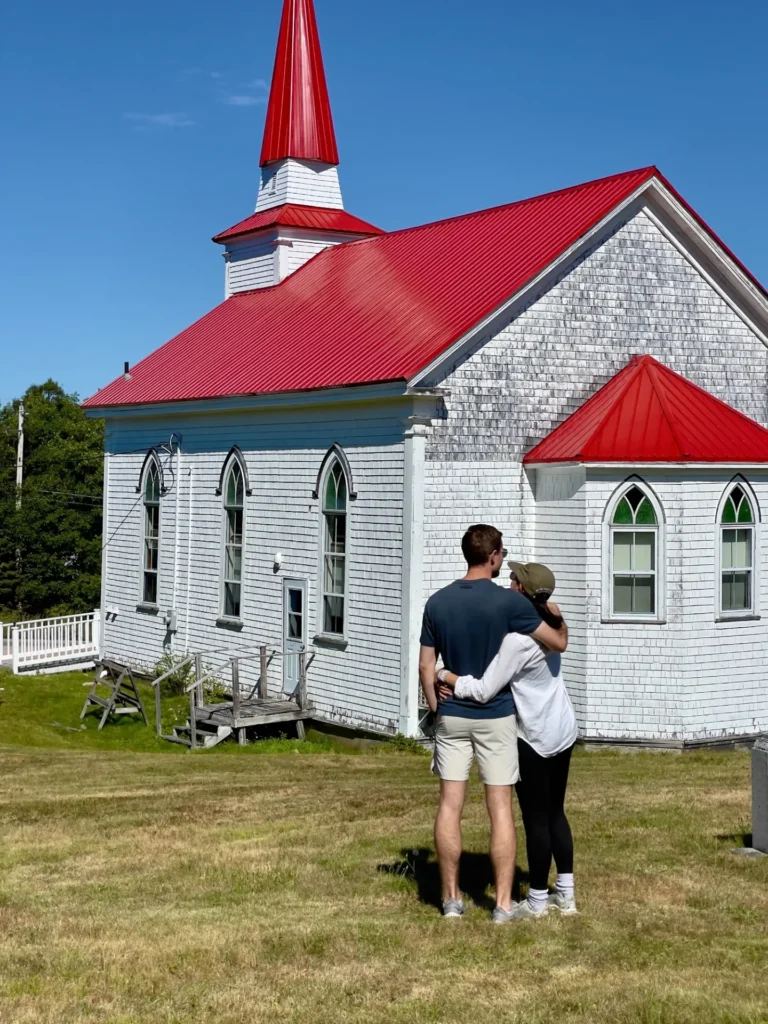 30’s couple embracing next to a white church with red roof in nova scotia, canada.