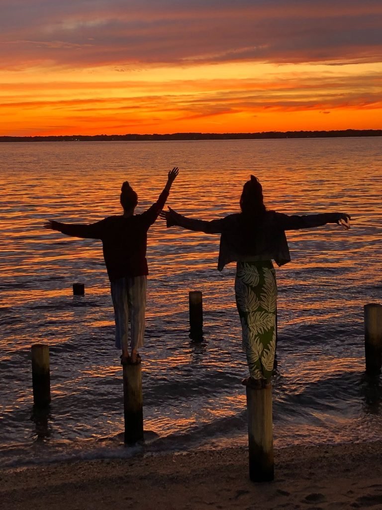 Two sisters balance on wooden posts along the shore as a vivid sunset colors the sky over shelter island, new york.