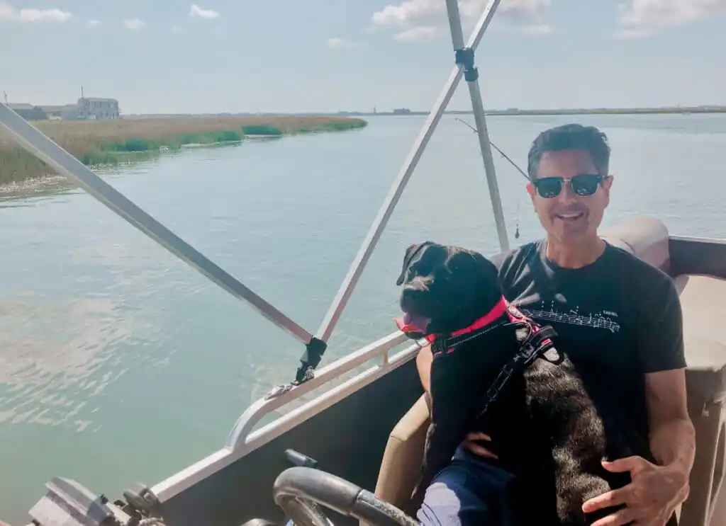 Man sitting on a pontoon boat holding a black dog on his lap, cruising along a calm river during a multigenerational family trip.