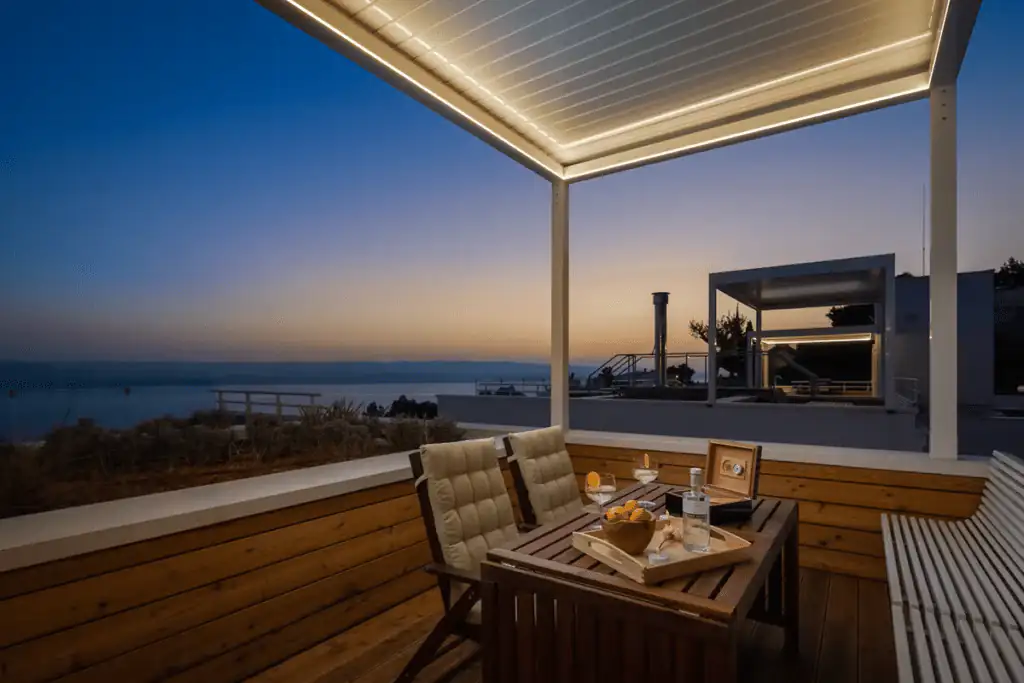 Covered terrace at dusk with a table set for drinks and snacks, overlooking the sea and a modern villa rooftop lounge.