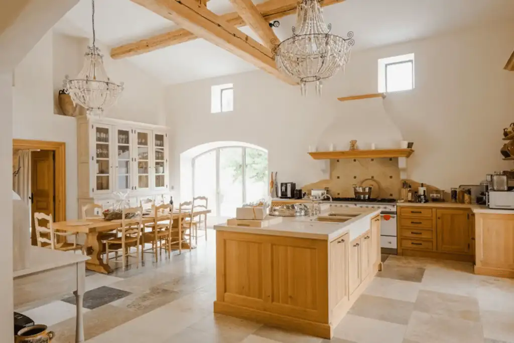 Bright country-style kitchen with exposed wooden beams, chandeliers, a large central island, and a dining table by glass doors.