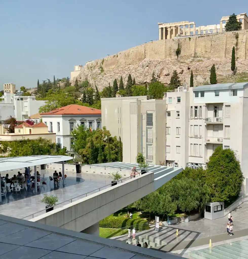 Original photo by paul partridge of an outdoor café terrace at the acropolis museum in athens, where visitors eat lunch with the parthenon visible on the hill above.