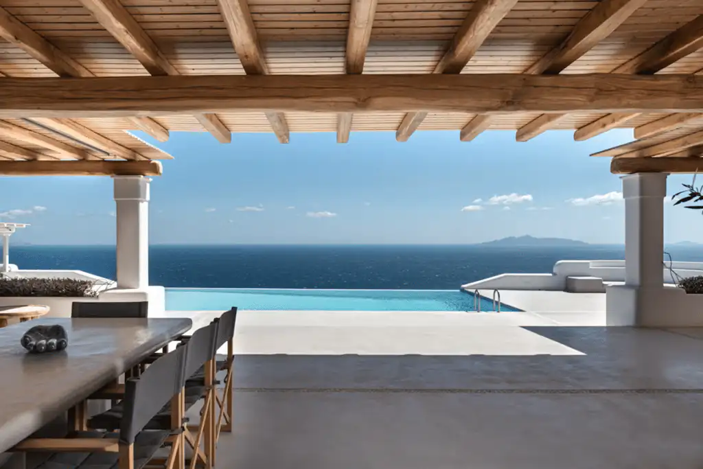 Shaded terrace dining area opens to an infinity pool and sweeping aegean sea views at a private villa in santorini, greece.