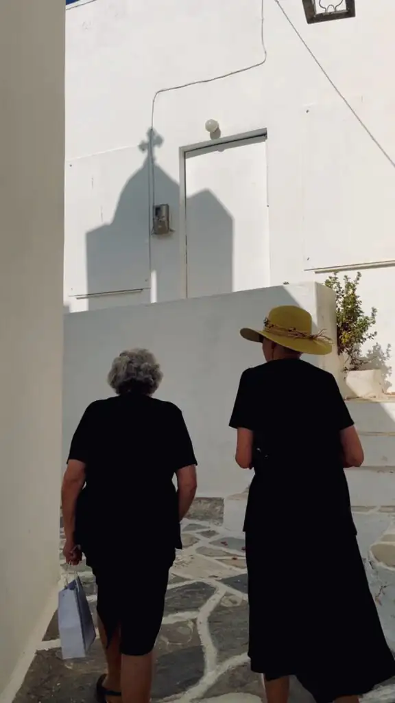 Original photo by paul partridge of two women walking along a whitewashed stone path in paros, greece, with a church shadow cast on the wall.