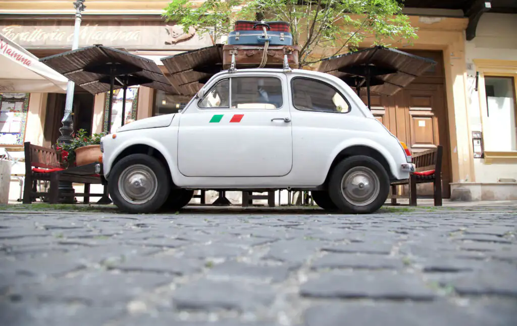 Tiny vintage white Italian car with luggage on top.
