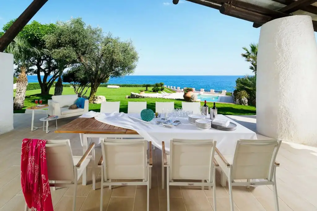 Outdoor dining area at Villa Oltremare in Sicily, overlooking a lush lawn, swimming pool, and the blue ocean beyond.