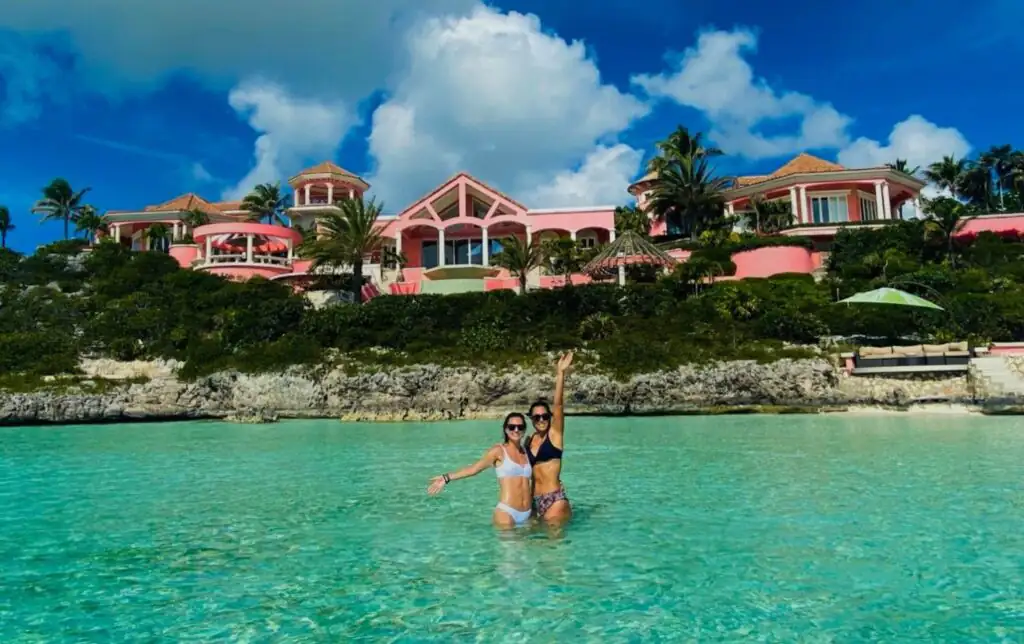 Two women stand in turquoise water in front of a pink waterfront villa in turks and caicos.